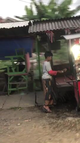 A person stands next to a red food cart. The cart is illuminated by overhead lights and displays various items, likely food or drinks, behind glass. The person is wearing a long skirt and a long-sleeved shirt. A sign with red and white branding is visible in the foreground. The background shows a building with a corrugated roof and some vegetation. The ground appears to be dirt or gravel.