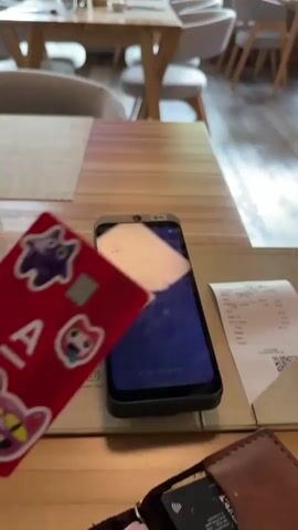 A person is paying for a bill at a restaurant. A phone on a wooden table is open to a payment app, displaying the price of 846 rubles. The person holds a red bank card, decorated with stickers, over the phone to make the payment. The phone then displays a swirling animation and the words "Жду ответа..." (Waiting for response). Finally, the screen turns a mix of green and blue as the payment goes through. A printed restaurant bill, a brown wallet, and other dining tables are visible in the background.