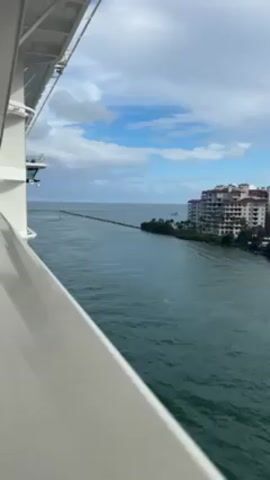 The video shows a view from the side of a cruise ship on a partially cloudy day. The camera is positioned towards the edge of the ship, with the white railing visible in the foreground. The view overlooks a body of water that leads out to the ocean. On the right side, there's a stretch of land with lush greenery and a tall, light-colored building complex. A narrow strip or barrier extends into the water, perpendicular to the shore. There appears to be a small boat or vessel in the water near the landmass. The sky is blue with scattered white clouds, and the sea is calm with a slightly greenish tint. The camera is stationary, and there are no observable people in the frame.