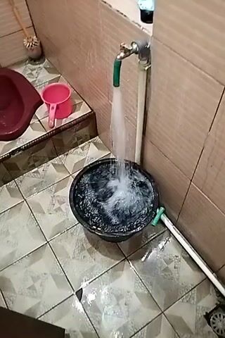A green hose is attached to a faucet and a stream of water is flowing from it into a large black bucket. The bucket is partially filled with water, and the water is splashing as it enters. The faucet is mounted on a white pipe, and there is a brown tiled wall behind it. To the left of the faucet, there is a pink plastic cup sitting on a ledge. On the floor, there are light-colored tiles with a geometric pattern. A long, thin white pipe runs along the floor from the bucket towards the right side of the frame, and there is a circular drain grate visible at the end of this pipe. The overall scene appears to be a bathroom or utility area.