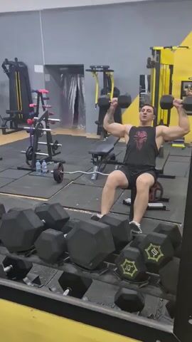 A man is sitting on a bench in a gym, performing dumbbell shoulder presses. He is wearing a black tank top and black shorts. There are various gym equipment visible in the background, including weight racks, benches, and machines. The gym has a gray and yellow color scheme. The man is holding a dumbbell in each hand, with his elbows bent and the dumbbells at shoulder level. He is extending his arms upwards, pressing the dumbbells overhead. The video is shot from a low angle, looking up at the man.
