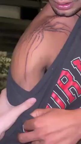 The video shows a man with a light brown skin tone and a tattoo on his shoulder. The tattoo is dark purple and appears to be a design with lines radiating outward. He is wearing a gray tank top with a large red and white design on it. A light-skinned hand is holding the tank top away from his body, possibly to display the tattoo more clearly. The background is blurry but appears to be an outdoor environment, possibly at night. There are some lights and trees visible in the background. The video is shot at a close angle, focusing on the man's shoulder and the tattoo.