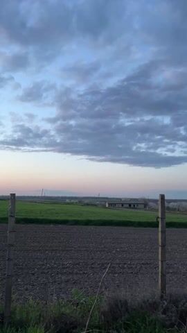 The video shows a landscape of fields under a cloudy sky. A freshly tilled field in the foreground transitions to a field of green crops in the midground. Behind these fields, a row of trees with white blossoms can be seen, along with a small brick structure with a dark, slanted roof.  A wooden fence runs along the edge of the foreground field. The sky is filled with clouds in shades of grey, with a hint of pink near the horizon.