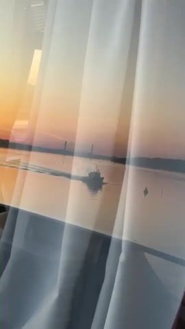 The video opens with a view through a window, showing a sunrise over a body of water. A small boat is moving across the water, leaving a wake. In the foreground, white curtains are visible, partially obscuring the view. The scene then shifts to a wider shot of the water and the sunrise, with what appears to be a city skyline in the distance. Another shot shows the deck of a boat with a railing and a wooden floor. A young person in a hooded jacket is standing at the railing, looking out at the water. The video concludes with a view from the boat of the water and the sunrise, with the railing of the boat in the foreground. The sky is a gradient of orange and yellow, and the water reflects the colors of the sunrise. There are a few small white specks in the distance that might be birds. A white buoy is visible in the water in one shot.