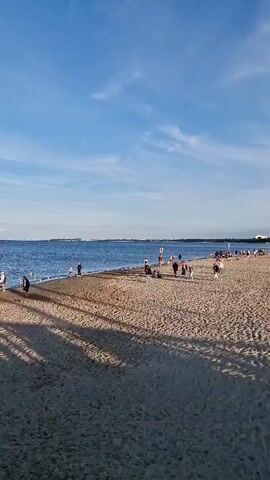 The video shows a beach scene with a calm blue ocean under a clear blue sky with some clouds. Several people are walking along the shoreline and on the sand. There are also some boats docked in the distance. Shadows of what appear to be trees or a structure are cast across the sand. In the foreground, there is a white railing and a flagpole with a white and blue banner. Two people are standing on the beach in the middle ground, facing away from the camera. The overall atmosphere is peaceful and serene.