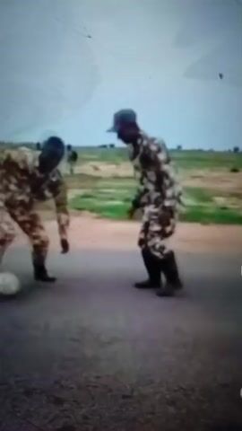 This video shows two individuals in camouflage uniforms and boots playing soccer. The scene appears to be outdoors, with a dirt road or path in the foreground and a field with sparse vegetation in the background. The sky is overcast. One individual is dribbling a soccer ball, while the other is positioned to receive a pass or intercept the ball. The overall impression is one of informal recreation amidst a possibly military or training environment.