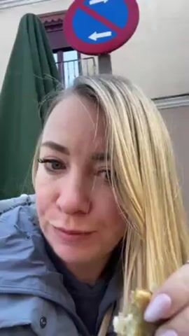 A woman with long blonde hair and wearing a grey jacket is eating a small piece of food outdoors. She looks directly at the camera as she takes a bite and chews.  Behind her is a green umbrella, a building, and a no parking sign with an arrow. She appears to be evaluating the taste of the food.