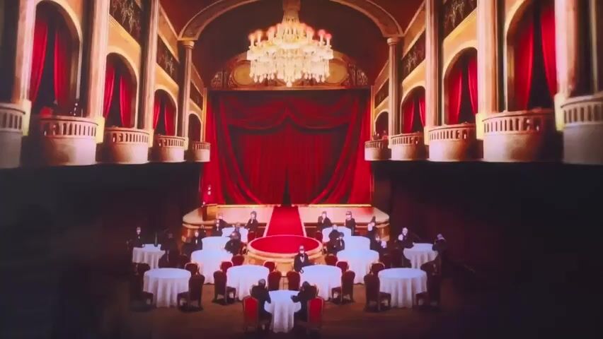 The media file depicts an ornate, dimly lit theater or ballroom. The main focal point is a stage draped with rich red velvet curtains and a crystal chandelier overhead, illuminated by soft, warm light. In the foreground, round tables draped with white tablecloths are set up, each seating several individuals. These individuals are dressed in dark formal attire and appear to be wearing masks, suggesting a masked ball or formal gathering. Some are seated, while others stand, possibly mingling or waiting.

The scene transitions to a close-up of a purple, stylized character with a glowing blue light on its head, resembling a unicorn or a mythical creature. This is followed by a shot of the same character, now appearing as a statue or a holographic projection, standing at the end of a red carpet leading towards a brightly lit, ethereal backdrop with red curtains framing the sides. The overall atmosphere suggests a formal event, perhaps a performance or a special occasion, with an element of mystery or fantasy introduced by the stylized character. The lighting is predominantly dramatic, using shadows and spotlights to create a sense of grandeur and anticipation. No explicit time of day or weather conditions are discernible, but the indoor setting implies a controlled environment. No visible text is present in the provided frames.