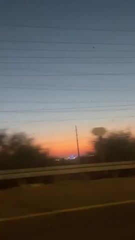 The video shows a blurry, moving view from inside a car at dusk or dawn. The sky transitions from a pale blue to orange and pink hues. Power lines stretch across the top of the frame. On the right side, a white car with bright headlights passes by. In the distance, there are trees and some lights, suggesting a city or town. The road has yellow lane markings. The interior of the car is dimly lit, with a dashboard visible and a red object in the lower right corner. The overall impression is that of a journey or commute as daylight fades.