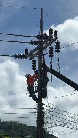 Two individuals in safety gear are working on a utility pole. They are both suspended from the pole using safety harnesses and ropes. One individual, wearing an orange top, appears to be adjusting something on the pole. The other individual, in a dark top, is positioned behind and slightly above the first, also working on the pole. The utility pole has multiple electrical wires running to it, supported by ceramic insulators. The sky is visible in the background with some clouds. The scene takes place outdoors, with trees visible in the distance at the bottom of the frame.