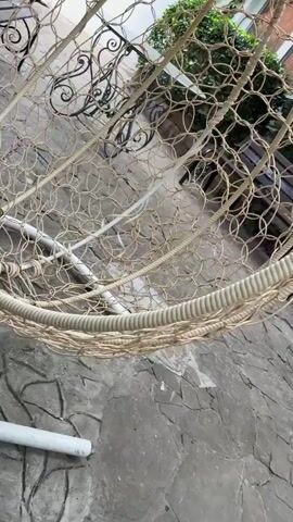 The video begins with a close-up of a woven hammock made of light-colored rope, revealing a textured, net-like pattern. The camera then pans right, showing a stone patio with irregularly shaped flagstones. In the background, a weathered wooden bench with black metal legs is visible. Beyond the bench, a building with white walls is adorned with lush green vines. A small window with a metal frame can be seen on the stone wall. Further to the right, a recessed niche in the stone wall holds three small, stylized figurines. The figurines appear to be angelic or robed figures, with one in black, one in gray, and one in white. They are arranged on a ledge within the niche. A red potted plant with green leaves is positioned in front of the stone wall, near the figurines. The camera continues to pan, showing a white door to the right of the niche and more greenery in the foreground.