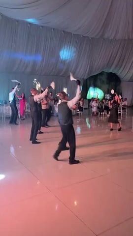 The video shows a group of men in formal vests and trousers dancing on a brightly lit floor, possibly a dance floor at a wedding reception or party. They are holding what appear to be sparklers or small fireworks in their outstretched arms, which are actively emitting sparks. The room is decorated with draped fabric overhead and lit with vibrant pink and red lighting, creating a festive atmosphere. In the background, other guests are seated at tables or standing, observing the performance. The men are moving in unison, suggesting a choreographed dance. One man at the far right is wearing a black dress and holding a white object, possibly a fan, and is also performing a dance.