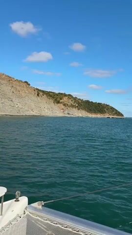 A video shows a scenic view of the sea and a rocky coastline from a boat. The water is a deep teal color with gentle waves. The coastline is characterized by a steep, rocky cliff face covered in sparse vegetation, primarily bushes and trees. There are a few small buildings nestled at the base of the cliff along the shoreline, appearing to be simple structures. The sky is a clear blue with a few white, fluffy clouds. The perspective suggests the video is being filmed from the front of a boat, with part of the boat's railing visible in the foreground. The overall impression is a serene coastal landscape on a bright, sunny day.