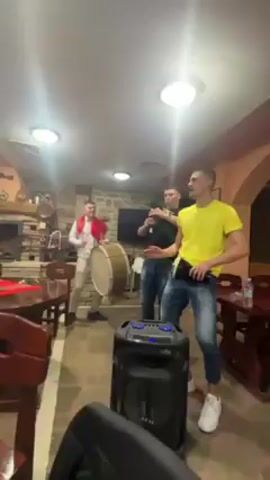 Three young men are enjoying themselves in a room that seems to be part of a restaurant or a home with a rustic feel. One man, dressed in a white shirt and light pants with a red scarf, is playing a large drum. Another man, wearing a dark shirt and jeans, is playing what appears to be a bagpipe. The third man, in a bright yellow shirt and ripped jeans, is dancing and playing a smaller drum. A large speaker sits in the foreground, likely providing music to which they are playing along. The room has stone walls, a fireplace with a roaring fire, wooden furniture, and is well-lit by ceiling lights. They appear to be having fun, energized by the music.