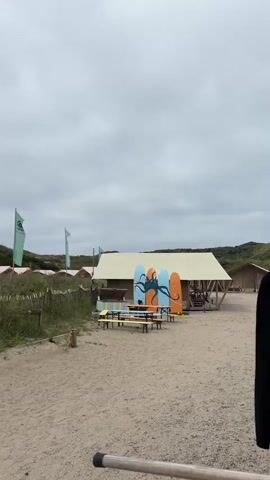 This video shows a beach campsite on a cloudy day. In the foreground, there is a wooden structure with horizontal bars. In the midground, there is a sandy area with picnic tables and benches, some of which are made of pallets. Behind the tables, there is a large tent structure and smaller buildings, possibly cabins. There are also several surfboards leaning against a structure, one decorated with a blue octopus. Green flags are visible in the background, and sand dunes covered in vegetation rise behind the campsite. The overall impression is of a relaxed, outdoor recreational area.