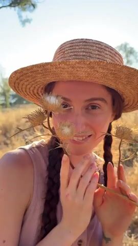The video shows a close-up of a young woman wearing a straw hat and a pink top. She has reddish-brown hair in two braids and is holding a dried thistle flower in front of her face. The background is blurred and appears to be an outdoor setting with dry vegetation and trees, suggesting a warm, sunny environment. The woman is looking at the camera and smiling in some frames, and her expression changes to a more serious or playful one in others. The thistle flower is positioned such that its spiky parts are close to her face, and she is holding it with her hands. The lighting is natural and seems to be from the sun.