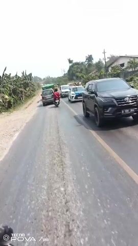 The video shows a street view from a moving vehicle. A single motorcyclist wearing a red shirt is seen from behind, traveling on the right side of the road. Several cars, including a white SUV with blue markings and a silver minivan, are also visible. The road is paved with a rougher shoulder on the left and bordered by vegetation on both sides. Utility poles with wires are present along the right side of the road, and houses are visible in the background. The sky is bright and hazy, suggesting daylight.