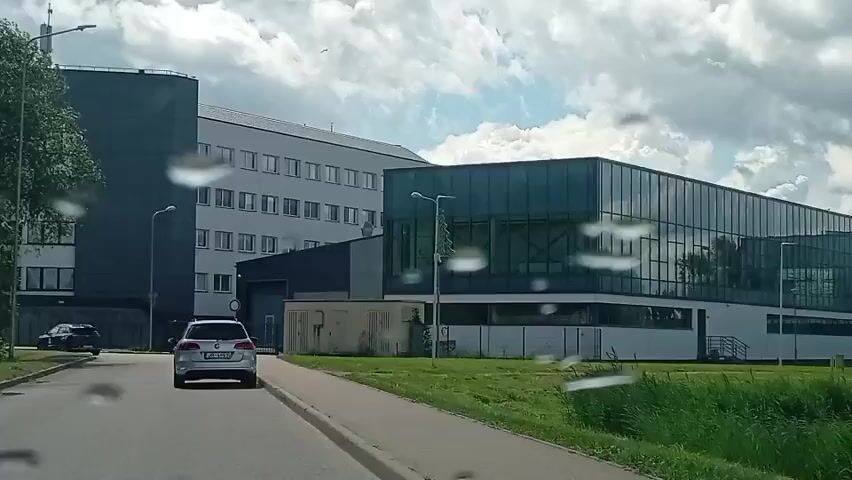 A dashcam recording shows a white car driving down a road. The car is in the left lane and is heading towards a modern building with large glass windows and a darker section. To the right of the building is a pond with some reeds. Further to the right, beyond the pond, there is a playground with a slide and other equipment. The sky is cloudy with patches of blue. The recording appears to be from inside a car, with raindrops visible on the windshield.