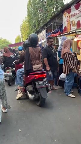The video shows a street scene with several people and vehicles, likely a market or bustling public area. In the foreground, a maroon scooter is stopped, with two people on it, both wearing helmets and modest clothing. The person in the back is wearing a brown outer garment and has a small purse slung over their shoulder. The scooter's license plate is visible: G 4891 ALB. To the left of the scooter, another scooter is partially visible with a rider wearing a brown hijab. Further to the left, a person with a patterned skirt and light blue top is walking away from the camera, also wearing a brown hijab. To the right of the scooter, a man in a black t-shirt and shorts is standing and looking towards the left. Behind him, more people are visible, including a woman in a hijab and a person in a striped top. In the background, there are stalls or shops with awnings, and trees line the street. The overall atmosphere appears to be daytime and crowded.