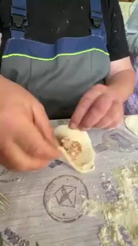 A person wearing gray overalls is shaping a piece of dough on a lavender-patterned tablecloth. Flour is scattered around the dough, and a fork lies to the left. The person's hands are visible, carefully folding and pinching the dough.