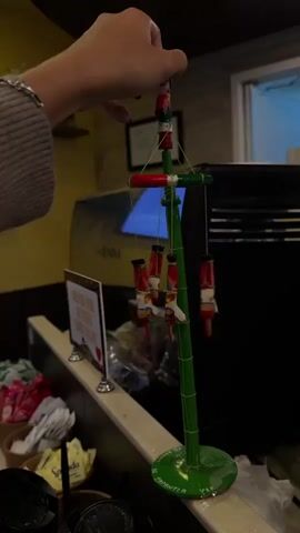 The video features a close-up shot of a person's hand interacting with a miniature "Voladores de Papantla" (Papantla Flyers) toy. The hand, with painted fingernails and a bracelet, initiates the spin of the vibrant green, red, and white cultural souvenir.

The toy consists of a central green pole with a spinning crossbar. Four small figures, adorned in traditional attire, are suspended by strings and rotate outwards, mimicking the traditional Danza de los Voladores ceremony. The green base of the toy is clearly inscribed with "PAPANTLA VER.", indicating its origin in Papantla, Veracruz, Mexico.

The scene is set indoors, likely within a cafe or coffee shop. In the background, a professional-grade coffee machine, partially displaying "LA ROMINA" on its surface, is visible. A counter runs along the right side, holding bowls filled with various condiment packets (such as sugar and sweeteners), and several disposable cups. A small sign on the counter, partially obscured, reads "GRACIAS POR SU VISITA" (Thank you for your visit). Overhead, a shelf with small decorative items is dimly visible. The lighting is artificial, illuminating the indoor space. No specific time of day or weather conditions are discernible.