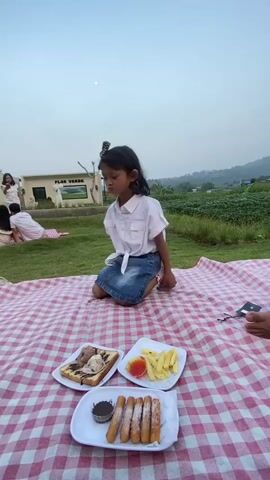 A young girl in a white short-sleeved shirt and denim skirt is kneeling on a red and white checkered picnic blanket. She is reaching her right hand out. In front of her on the blanket are three plates of food: one with toast and ice cream, one with french fries and ketchup, and one with churros and chocolate sauce. In the background, other people are picnicking on the grass, and there are buildings and trees. The sky is overcast.