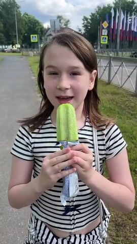 A young girl with brown hair stands on a sidewalk, holding a green popsicle. She wears a black and white striped t-shirt and a silver purse is slung across her body. The girl is smiling and about to take a bite of the popsicle.

The background shows a grassy area with a fence and a building. Flags are visible in the distance. The sky is cloudy, suggesting it is daytime. The scene appears to be in Boksitogorsk, Russia. The girl's expression is happy and she seems to be enjoying her treat. The popsicle is light green and appears to be covered in a fine white powder, possibly sugar or frost. The wrapper of the popsicle has some visible text, but it is too blurry to read.
