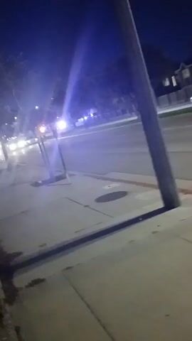 The video is shot at night from a sidewalk, looking out onto a street. There are some cars in the distance, and a vehicle with flashing lights is closer. There's also a street lamp and other indistinct objects further up the street. The sidewalk is pale gray, and there is a pole between the camera and the street.