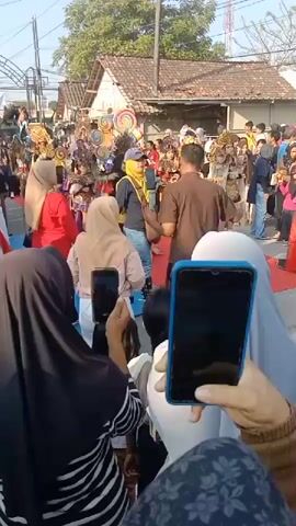 The video shows an outdoor street event with a parade. In the foreground, several people are holding up smartphones to record the event. Two smartphones are prominently visible, one in blue and one in black. The people in the foreground are wearing various headscarves. In the middle ground, there is a red carpet laid out on the street. Several individuals in colorful, ornate costumes are walking on the carpet. One individual in the center wears a yellow garment with elaborate headwear. Other participants in the parade have similar elaborate costumes and headdresses. The background shows houses lining the street and a clear sky, indicating it is daytime. There are also spectators in the background watching the parade. The overall scene suggests a festive community event or cultural celebration.