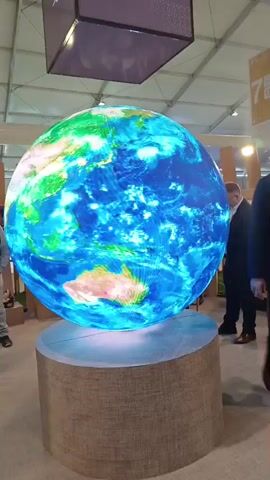 This video captures an indoor scene, likely an exhibition or convention in Al Machouar – Stinia, Morocco, centered around a large, spherical LED display. The display, mounted on a textured cylindrical base, continuously rotates, showcasing a vibrant, animated view of Earth with continents and oceans.

Throughout the video, various national flags are superimposed onto their respective countries on the globe: the US flag appears over North America, followed by the Iraq flag over the Middle East, and then flags for Saudi Arabia, the UAE, and Qatar also in the Middle East region.

Several people, mostly dressed in business attire, are visible in the background and walking past the display, observing it. The setting features a high, tent-like ceiling structure and general bright indoor lighting. Muffled conversations can be heard in the background.