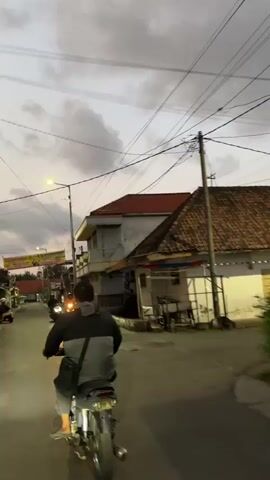 The video begins with a rear view of a person riding a motorcycle down a street. The rider is wearing a dark jacket with a gray stripe across the back and a bag slung over their shoulder. The motorcycle is a mid-size model with a visible exhaust pipe. The street appears to be in a residential area with houses on either side. The houses have tiled roofs and are of varying architectural styles. Overhead, there are numerous power lines crisscrossing the sky. The sky itself is overcast with a twilight hue, suggesting either early morning or late evening. Several other motorcycles are visible further down the street, also in motion. On the left side of the street, a banner with text is hanging from a pole. The road surface is paved and appears to be wet in some areas, possibly indicating recent rain. The overall atmosphere is subdued, consistent with the low light conditions.