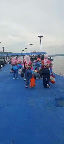 A group of people are standing and walking on a blue pier. They are wearing matching outfits, long-sleeved shirts with colorful patterns and jeans. Some are wearing headscarves. They are carrying bags and appear to be on a trip or excursion. In the background, there is water, boats, and a distant shoreline with hills under an overcast sky. The pier has lampposts.