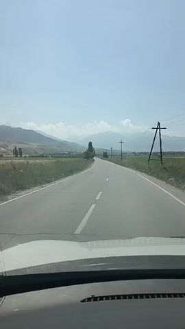 The video is a first-person perspective from inside a vehicle traveling on a paved road. The road has a white dashed line down the center and is bordered by dry grass and some trees on either side. In the distance, there are mountains under a clear blue sky with some scattered clouds. Telephone poles and power lines are visible along the side of the road. The vehicle appears to be moving at a moderate speed.