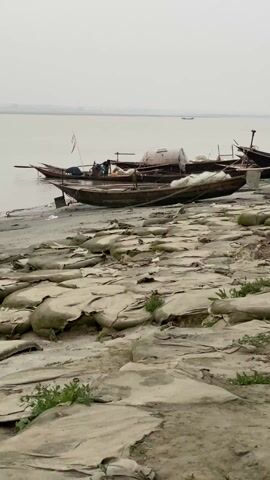 The video shows a river bank on a gray, overcast day. Several wooden boats are beached along the shore. The river water is a dull gray color, merging with the hazy sky in the distance. The foreground consists of a sandy area covered in numerous large, flat, gray objects, resembling hides or tarps, scattered across the surface. There are sparse patches of green vegetation emerging between the hides. A man can be seen in one of the boats. The only sound is a recording of Azaan, a Muslim call to prayer.