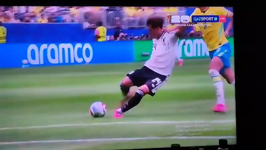 The video shows a soccer match. A player in a white and black uniform is attempting to kick a soccer ball. A player in a yellow uniform is in the background. Another player in a purple uniform dives to save the ball. The soccer ball is on the green field, near a white goal line. The background shows a stadium with spectators. The QAZSPORT logo is visible in the upper right corner of the screen. The word "aramco" is visible on a blue banner in the background.