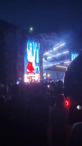 A large outdoor concert is taking place at night. A crowd of people is gathered, facing a stage where a performance is happening. A large LED screen displays a woman singing, with text below her. Other performers are visible on stage. The lighting is dramatic, with blue and white spotlights illuminating the stage and the performers. Smoke or fog is present in the air. The overall atmosphere is energetic and festive.