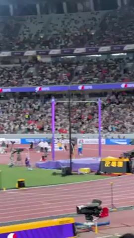 The video shows a pole vault athlete clearing the bar at a stadium event. The athlete, wearing a yellow and blue uniform, vaults over the bar, landing safely on the cushioned mat. A large crowd is visible in the stands, cheering enthusiastically. A person in the foreground raises their fist in celebration. The environment is a well-lit stadium with timing equipment and sponsor logos visible.