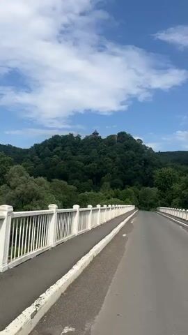 This image captures a scenic view from a bridge in Kamianytsia, Ukraine, on a bright, sunny day. The bridge, with its distinctive white railings, stretches forward, offering a path for vehicles on its asphalt surface. In the distance, a prominent hill covered in lush green trees dominates the landscape. Perched atop this verdant hill, a structure, possibly a castle or a large building, is visible, adding a historical or architectural element to the natural setting. The sky above is a vibrant blue dotted with scattered white clouds, indicating fair weather. The scene exudes a sense of tranquility and offers a picturesque view of the Ukrainian countryside.