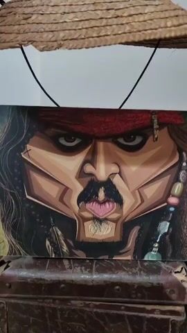A close-up shot shows a caricature of Jack Sparrow's face. A straw hat is placed on top of the artwork, obscuring the very top of the image. A hand is seen adjusting the hat, then lifts it off the artwork. The artwork is positioned on top of a brown chest. The background is blurry, showing a bookshelf with books.