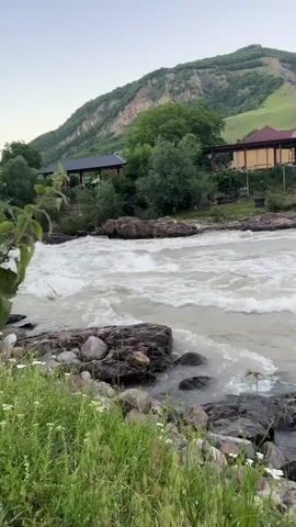 The video showcases a rushing river flowing through a mountainous landscape. The water appears murky, with visible rapids and whitecaps indicating a strong current. Rocks are scattered along the riverbed and on the banks, some submerged in the water and others forming small islands.

In the background, there are several buildings situated on a hillside overlooking the river. These appear to be houses or possibly some sort of establishment, with a few trees and green vegetation surrounding them. The landscape is predominantly hilly, with one prominent mountain in the distance. The slopes of the hills are covered in green grass, suggesting a rural or semi-rural setting. A cluster of trees is visible between the river and the buildings.

The time of day appears to be late afternoon or early evening, as the sky is a pale, clear hue, and the light is soft. The weather seems pleasant, with no indication of rain or strong winds. The overall mood is tranquil and natural, highlighting the power of the river and the serenity of the surrounding environment. The location is identified as Каменномостское сельское поселение, Russia. There is no visible text in the media file.