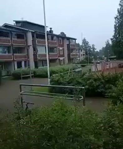 The video shows a residential area with apartment buildings and a playground. The ground is flooded, with water covering most of the visible area. Bushes and trees are present throughout the scene. A swing set and a small playhouse are visible in the playground. The sky is overcast, suggesting rainy weather. There are no people visible in the footage.