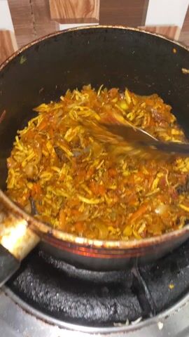 A pan of food is being stirred with a yellow spatula. The food appears to be a mixture of shredded chicken, onions, and carrots, seasoned with a yellow spice, likely turmeric. The pan is black and sits on a gas stove, which is turned on. The background features a tiled wall with a light brown and white pattern.