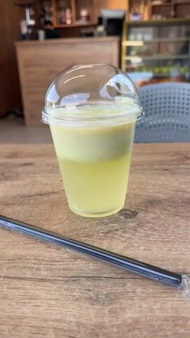 A close-up shot focuses on a clear plastic cup filled with a yellow, frothy beverage, topped with a clear dome lid. A black reusable straw lies diagonally across a wooden table in the foreground. A hand reaches into the frame, grasping the cup, and lifts it slightly. The background shows a blurry indoor cafe setting with a counter and display cases, transitioning to an outdoor view through a glass door, revealing a green lawn and trees under a bright sky.
