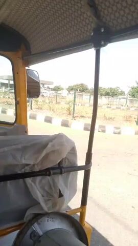 From the perspective of someone inside a moving three-wheeled vehicle, possibly a tuk-tuk or auto-rickshaw, the video shows a view of a road and the surrounding landscape. A gas station with a tall price sign is visible across the street, behind a chain-link fence. The sky is clear and bright. The vehicle’s interior frame and roof are visible, including some covered objects in the passenger area. The road has a black and white curb.