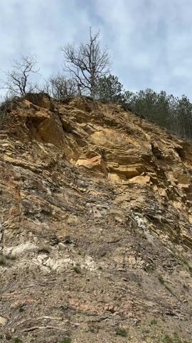 The video shows a dirt road winding through a valley. The road is surrounded by rocky hills covered in trees. The sky is cloudy and overcast. The rock formations on the hillsides are layered and vary in color from brown to yellow. The camera pans across the scene, showing the different textures and colors of the landscape.