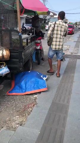 The video shows a street scene. On the left, a man in a plaid shirt and shorts stands near a fruit cart. He is facing a person on a red motorcycle, who is wearing a helmet and dark clothing. In the background, a cement truck drives down the street, and a red car is visible behind it. To the right of the motorcycle is a blue scale on the sidewalk. The ground is paved with concrete slabs and some dirt. A blue tarp is spread out on the ground near the fruit cart. The sky is overcast.