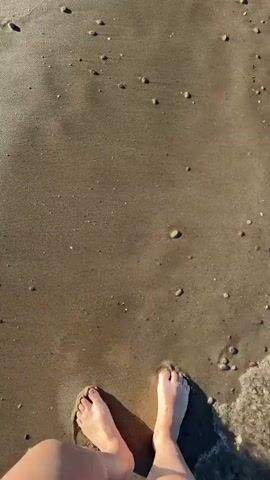A low-angle video shows a person walking barefoot on a sandy beach. The person's fair-skinned legs and feet are visible, with their shadow trailing behind them. The wet sand is a mix of light and dark brown, scattered with small pebbles. Clear ocean water, edged with white foam, washes over the sand near the edge of the frame. The lighting suggests a sunny day, highlighting the texture of the sand and the movement of the water.