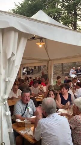 The video shows a group of people seated at long wooden tables under a white tent. The tables are set with plates, glasses, and bottles. People are interacting with each other, talking, and eating. In the background, there are trees and foliage, suggesting an outdoor setting. The lighting is dim, indicating it might be evening. Some individuals are looking at the camera, while others are engaged in conversation. A man in the foreground appears to be gesturing with his hands as he speaks. The overall atmosphere seems to be a social gathering or a meal.