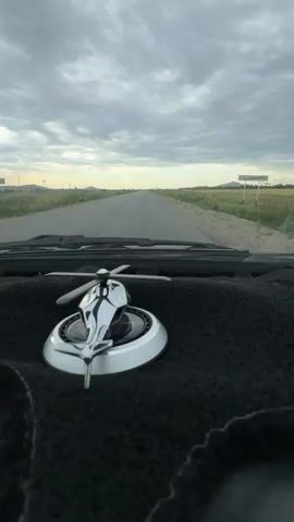 This is a first-person perspective video taken from inside a vehicle. The camera is focused on the dashboard, which is covered in black fuzzy material. A shiny silver helicopter-shaped ornament is prominently displayed on the dashboard, with its rotors slightly blurred, suggesting it may be moving due to the car's motion or air currents. Beyond the dashboard, the view through the windshield shows a paved road stretching into the distance. The road is flanked by fields of green and yellow vegetation, possibly sunflowers, under a cloudy sky. The horizon is marked by distant hills or mountains. The overall impression is of a road trip through a rural landscape.