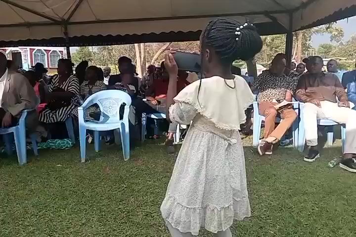 A young girl in a patterned dress and a white ruffled collar stands in the foreground facing away from the camera, holding a smartphone horizontally to record something off-screen. She has braided hair styled in a bun. Behind her, an outdoor gathering takes place under a large white tent. Several rows of chairs are set up on a grassy area, occupied by a diverse group of adults. Some individuals are looking towards the girl, while others are looking towards the left side of the frame where a woman in a patterned dress and a man in a suit are standing near a keyboard and audio equipment. The atmosphere appears to be a public event or celebration. The lighting suggests daytime with natural sunlight.